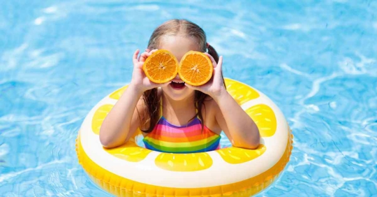 A young girl in a pool with a citrus hoop around her. She is holding up two halves of an orange, one in front of each eye to look silly