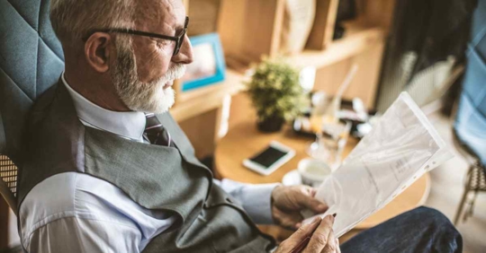 An elderly man sitting on a chair. He holds both a pen and a piece of paper that he is making notes on