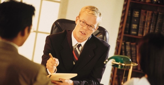 An attorney sitting behind his desk taking notes while a prospective client shares their story.
