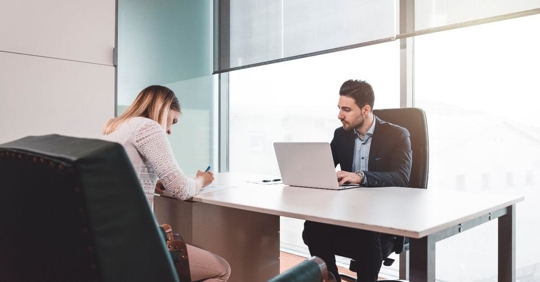 A woman sitting across the desk from a male attorney. They are working on something.