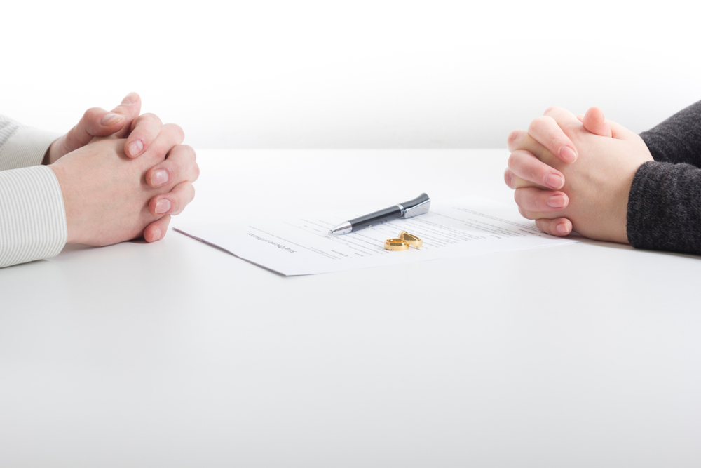 Couple sitting on opposite sides of a table with a paper, pen, and wedding bands between them.