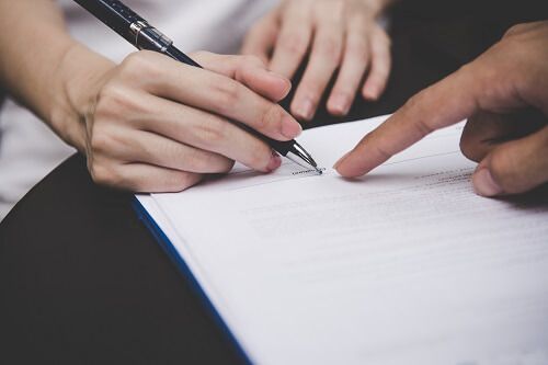 Close up of a hand holding a pen on a document while another hand points at the signature line.